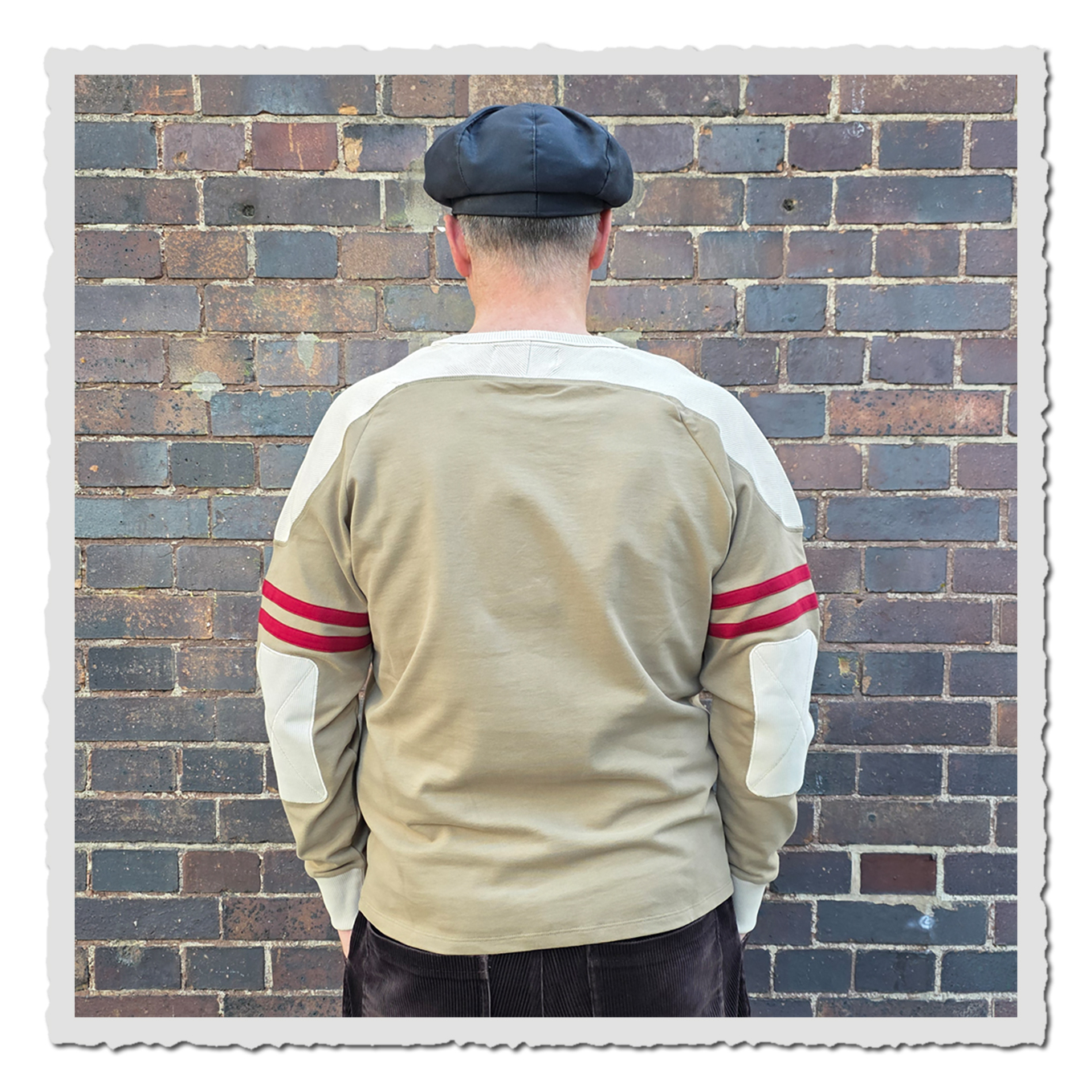 Rückansicht einer Person mit beigem Langarmshirt, hellen Schulterpartien und roten Ärmelstreifen.